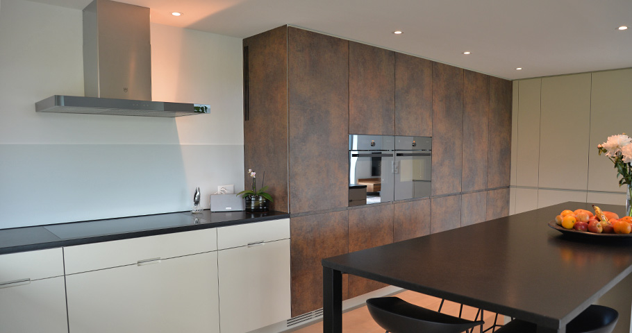 Küche mit schwarzen Esstisch, dunkler Arbeitsfläche dahinter, darunter helle Schrankfronten. Rechts hohe Wandschränke mit Kunstharz belegt
